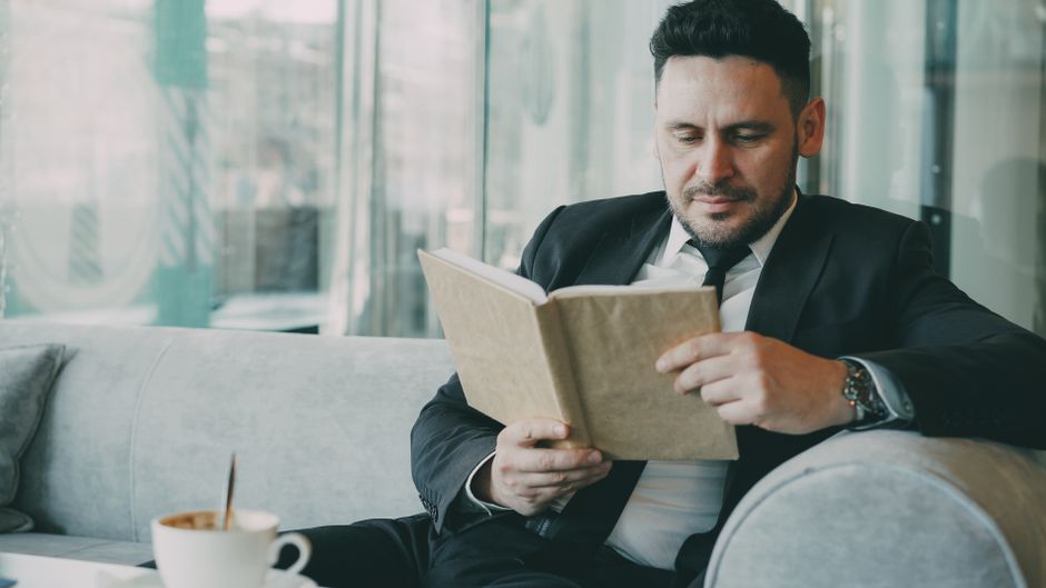 A businessman in a suit reads a book while relaxing in a cafe setting.